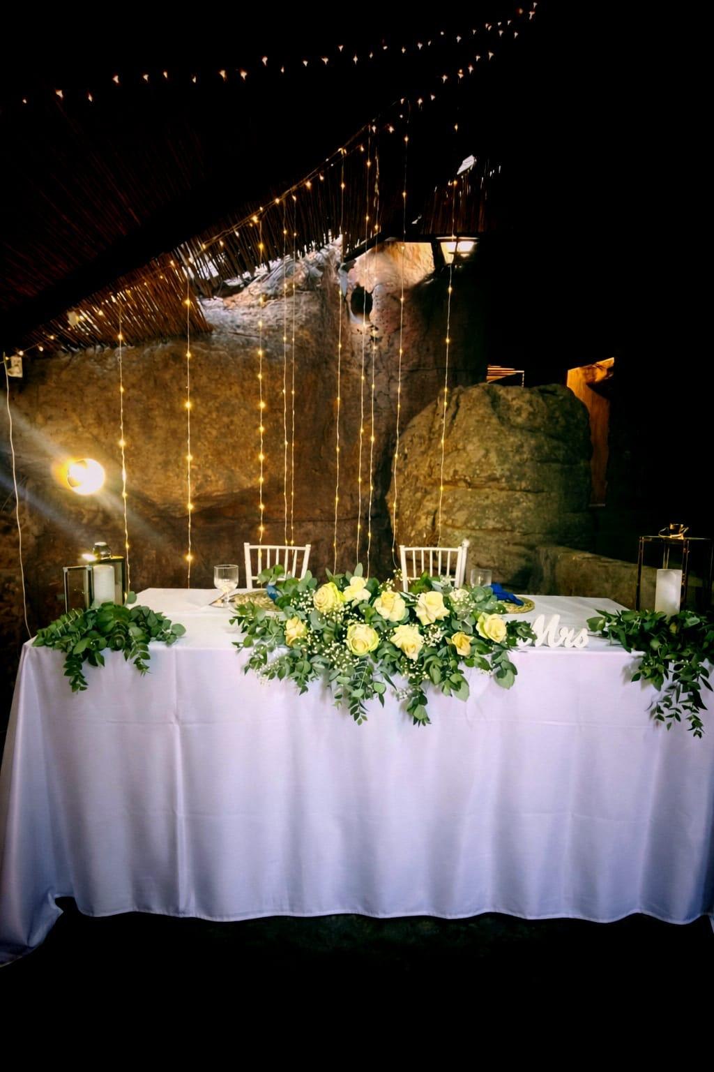 Wedding sweetheart table decorated with white roses and fairy lights against the rock wall at Donkieskraal